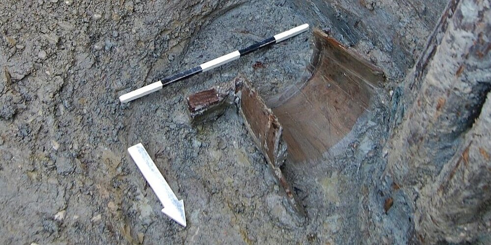 Daubeneimer in feuchter Erde in einem holzverschalten Schacht. In der Ecke steht eine Fototafel, vor dem Objekt Maßstab und Nordpfeil.