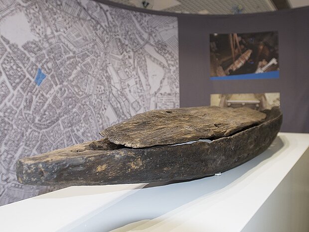 Ein längliches, bootsförmiges Holzobjekt mit Löchern und Deckeln steht auf einem Podest vor einem Stadtplan im MVG-Kundencenter.