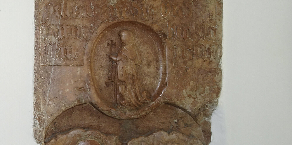 Grabstein aus rötlichem Marmor mit Darstellung einer Nonne mit Kreuz und Rosenkranz in rundem Fenster. Darunter befinden sich Wappendarstellungen, auf dem übrigen Stein eine Inschrift.