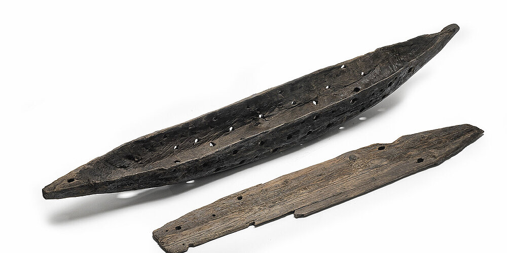 Längliches, bootsartiges Holzobjekt mit Löchern und daneben liegendem Deckel aus Schacht 5 am Marienhof.