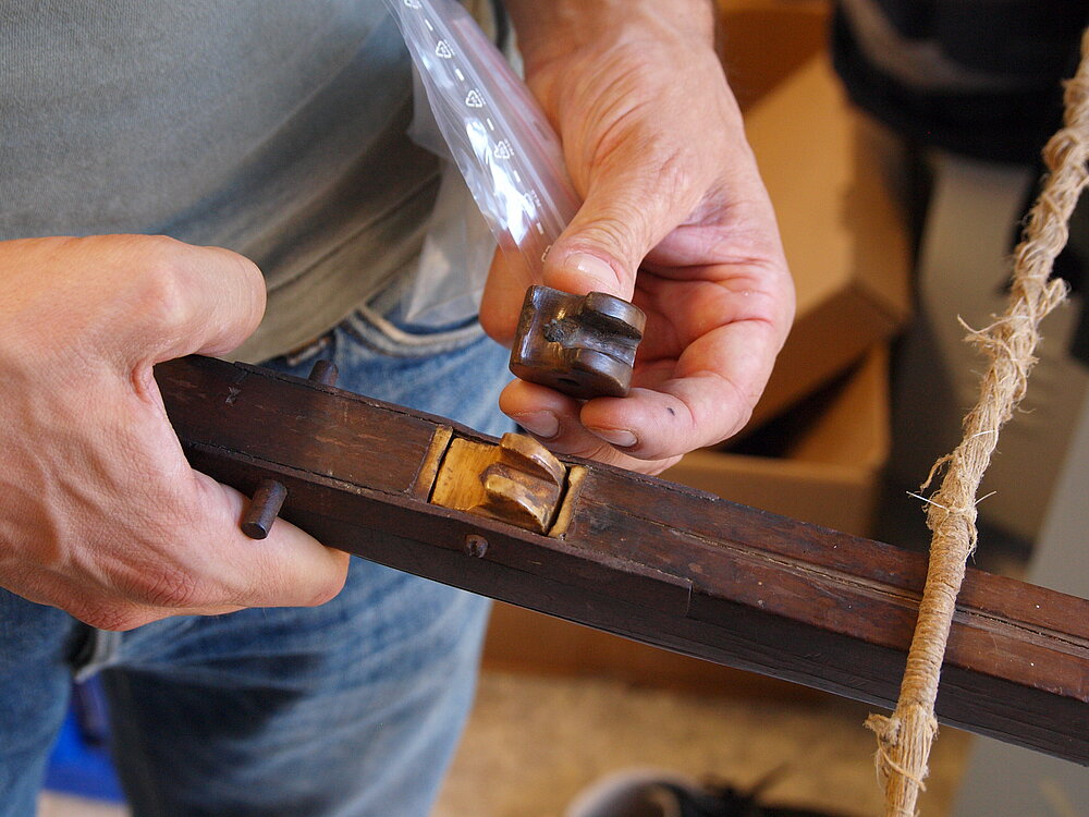 Ein Mann hält eine Armbrust in einer Hand, in deren Schaft eine Armbrustnuss eingelegt ist. In der anderen Hand hält er ein originales Fundstück aus dem 16. Jahrhundert neben die Armbrust.